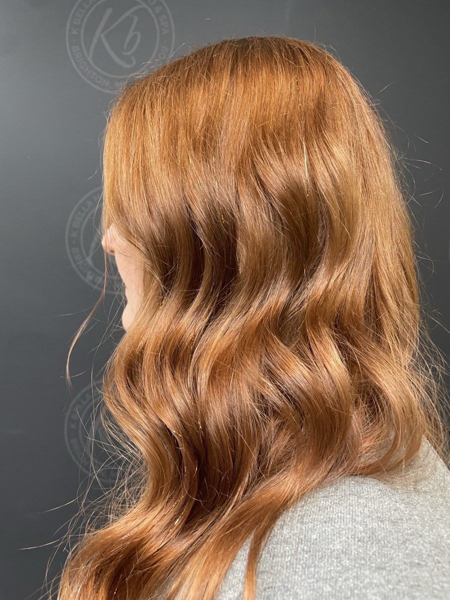 Back view of long wavy hair in warm caramel tones with tinsel accents, showing feathered layers and movement by Kristan Sayers at K Bella Hair Studio & Spa in Brighton, MI