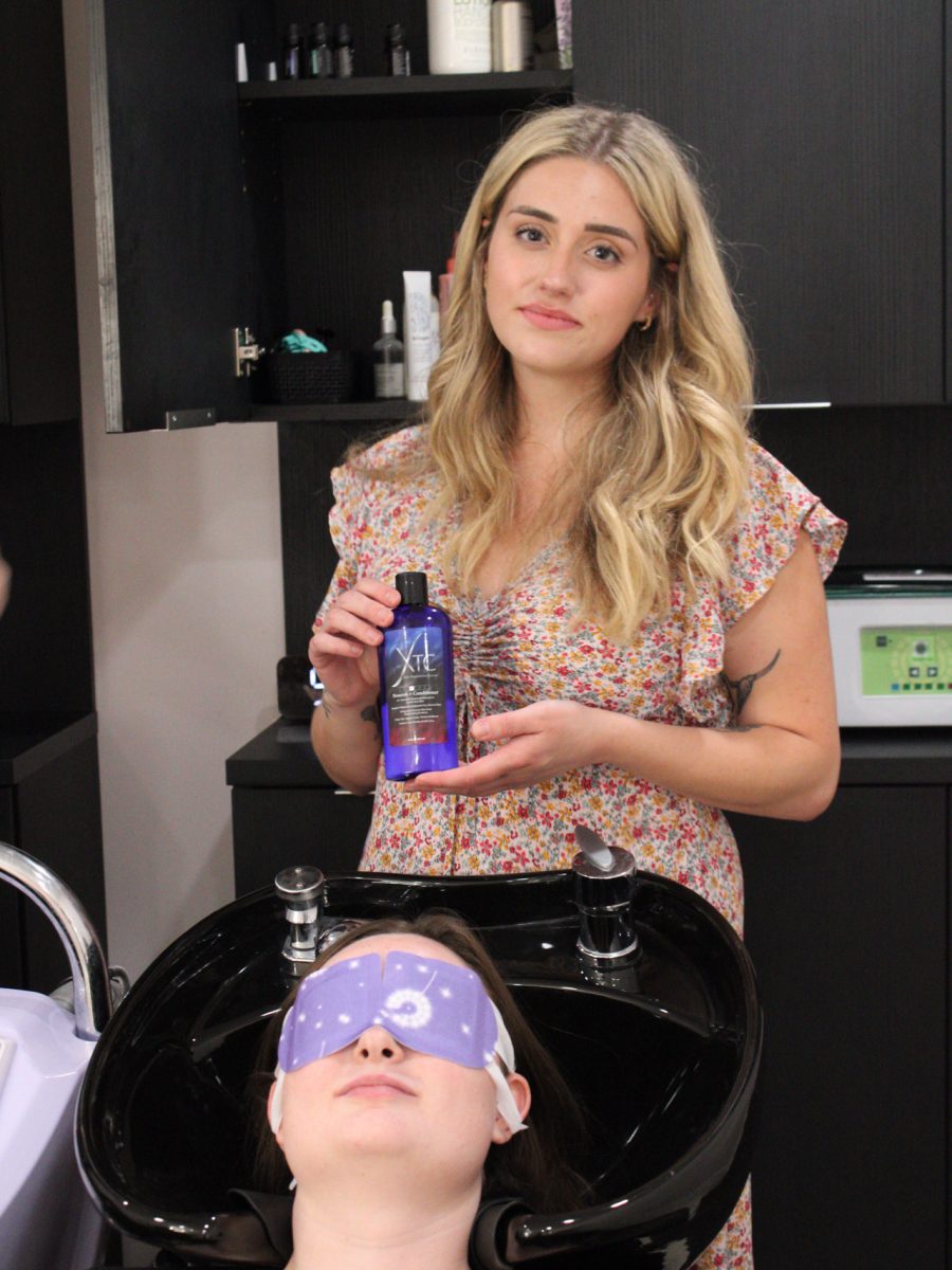 Client reclines with a purple treatment mask on her scalp while stylist holds a product bottle by Liberty Greenwood at K Bella Hair Studio & Spa in Brighton, MI