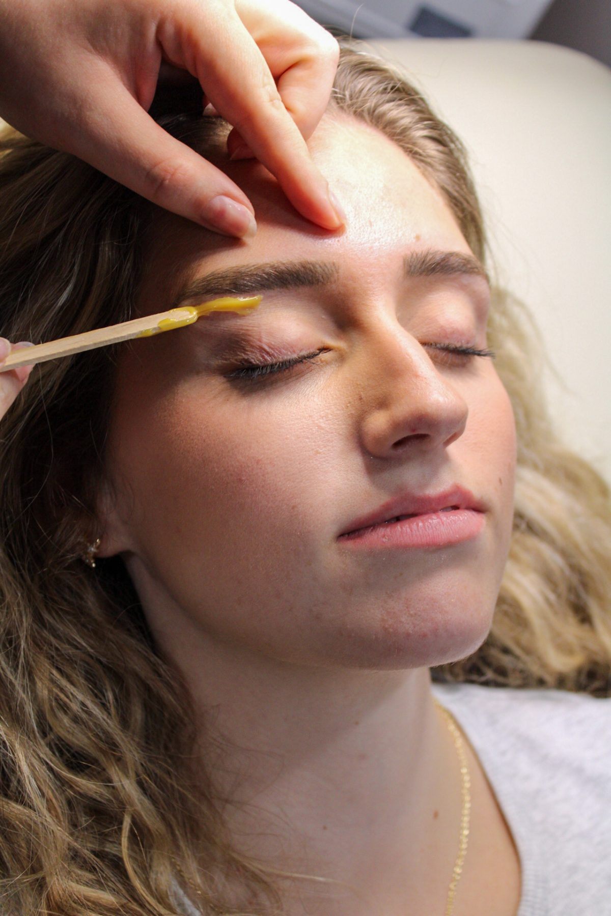 Esthetician applying warm wax with a spatula to shape a client's eyebrow at K Bella Hair Studio in Brighton, Michigan